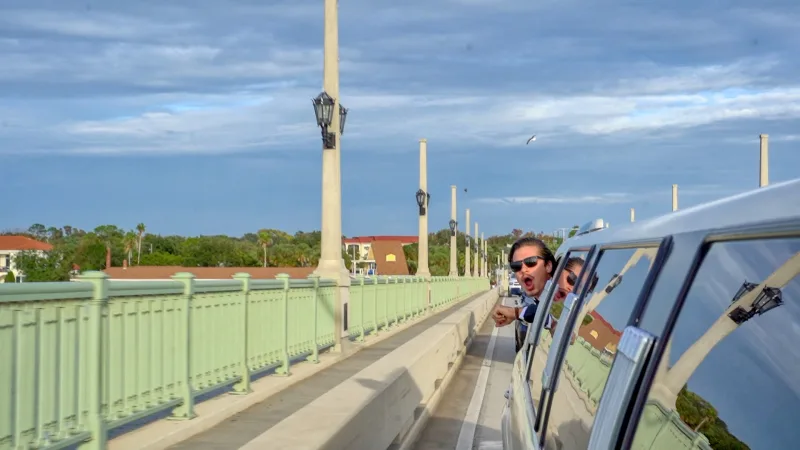 A grinning groom in sunglasses leaning out of a white limousine window as it crosses the Bridge of Lions, with the bride beside him