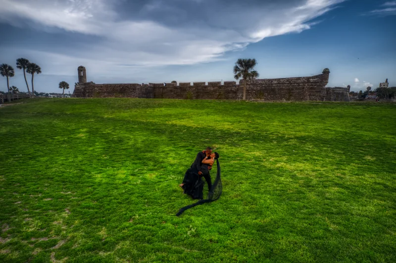 A couple embracing on the green lawn in front of Castillo de San Marcos, the bride in a black gown with a long black veil trailing across the grass