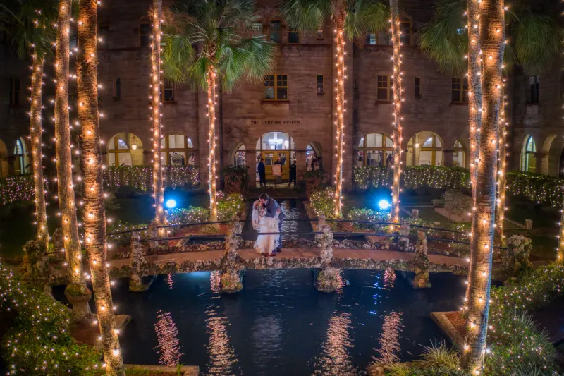 A bride and groom kissing on the small stone bridge in the Lightner Museum courtyard at night, the palm trunks wrapped in warm string lights and reflected in the pond below