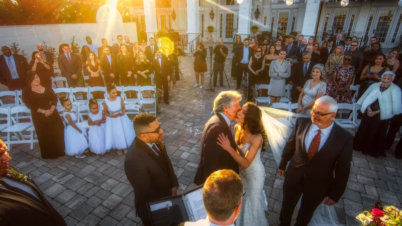 An outdoor ceremony at River House in St. Augustine, filmed from above at sunset: the bride receives a kiss at the altar while flower girls in white dresses and standing guests look on
