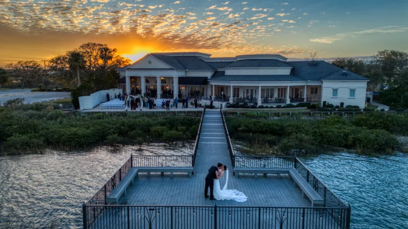 Drone shot at sunset of a couple kissing at the end of the dock at River House in St. Augustine, the bride's long veil spread across the boards and wedding guests gathered at the white columned building behind them