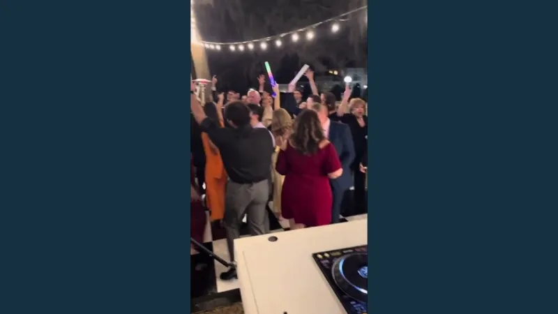 Guests with arms raised and glow sticks on a black and white checkered dance floor under string lights at night, seen from behind the DJ's turntable