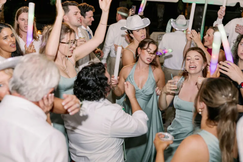 Bridesmaids in sage green dresses and guests in white cowboy hats waving foam glow sticks on a crowded dance floor at the Fountain of Youth