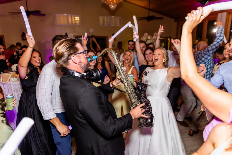 COS Celebrations founder Corey Peterson playing saxophone in the middle of a packed dance floor at the Fountain of Youth, the bride beside him and guests waving foam glow sticks