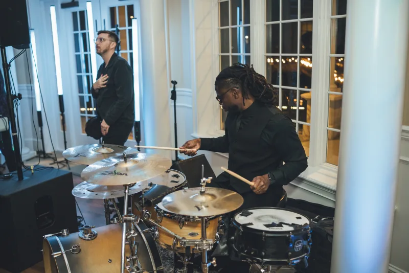 A drummer at a full kit playing beside the DJ setup at River House, tall white-framed windows and blue uplighting behind him and a second musician standing to the left