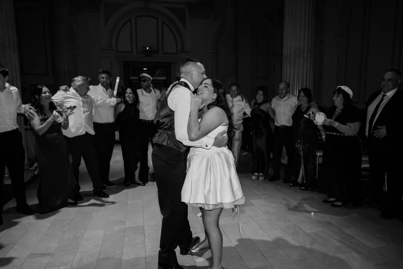 Black and white photo of a bride and groom kissing on the dance floor at Treasury on the Plaza with guests gathered in a ring around them