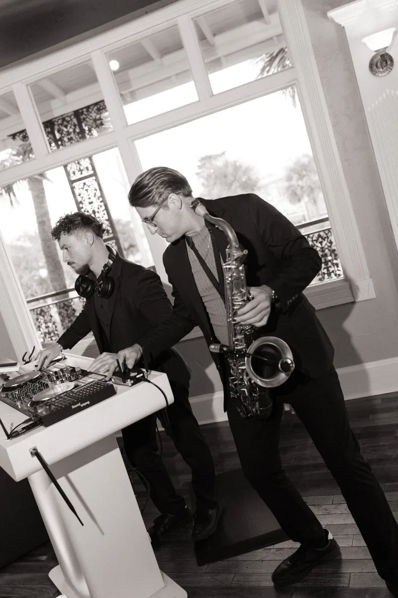 Sepia-toned photo of a saxophonist playing beside the DJ at a white booth in front of the tall windows at The White Room, the DJ working the decks with headphones around his neck