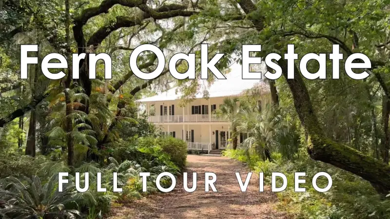 A white two-storey house with a wraparound porch at the end of a gravel drive, under a canopy of oaks, palms and ferns, with the words Fern Oak Estate Full Tour Video overlaid