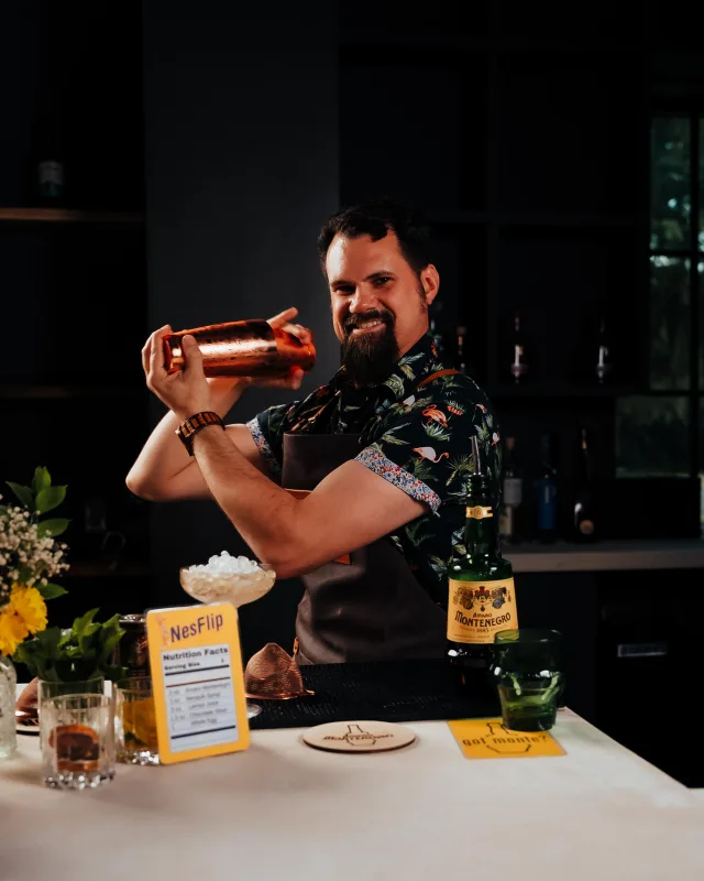 A bartender in a floral shirt and leather apron smiling as he shakes a copper cocktail shaker behind a bar, with a bottle of Amaro Montenegro, a bowl of crushed ice and a hand-drawn recipe card set out in front of him
