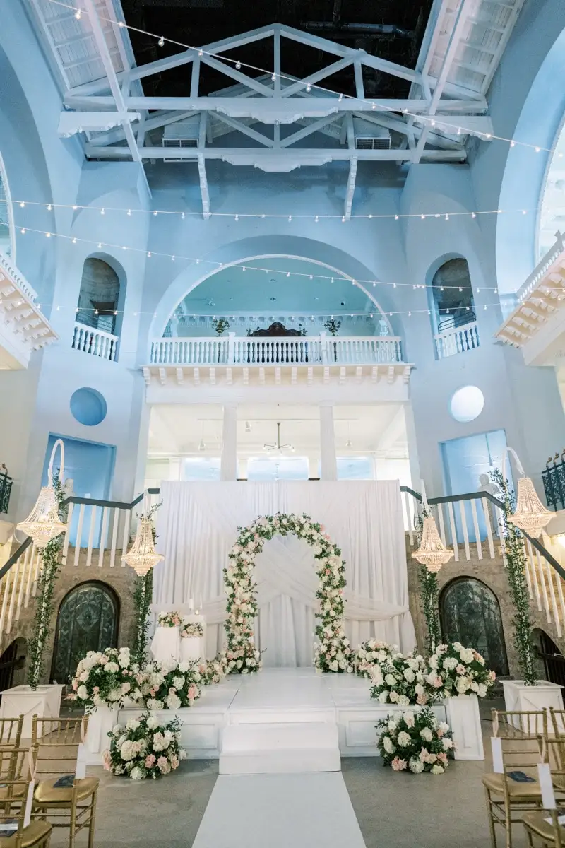 Ceremony setup at the Lightner Museum in St. Augustine: a pink and white rose arch on a white stage, rose arrangements along its front, greenery climbing the crystal chandelier stands, and string lights across the tall courtyard