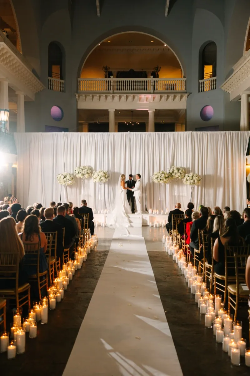An evening ceremony at the Lightner Museum, the aisle lined on both sides with pillar candles and tall white floral arrangements on stands flanking the couple against a white draped backdrop