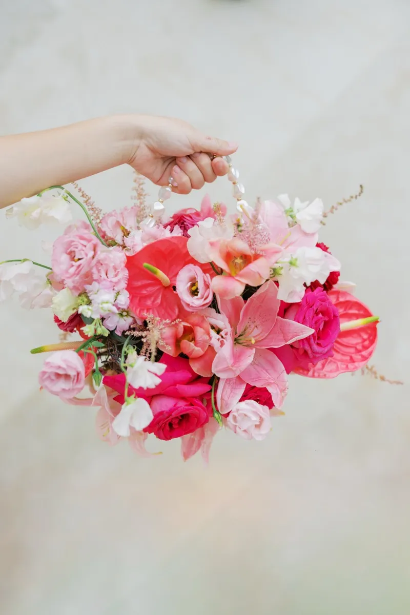 A hand holding a hot pink and blush bouquet of anthurium, lilies, garden roses and sweet pea by a strand of pearls