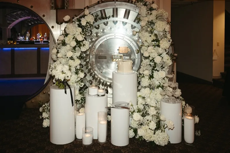 White roses, hydrangea and baby's breath framing the open bank vault door at Treasury on the Plaza, with a white cake, a stack of champagne coupes and pillar candles on white columns in front