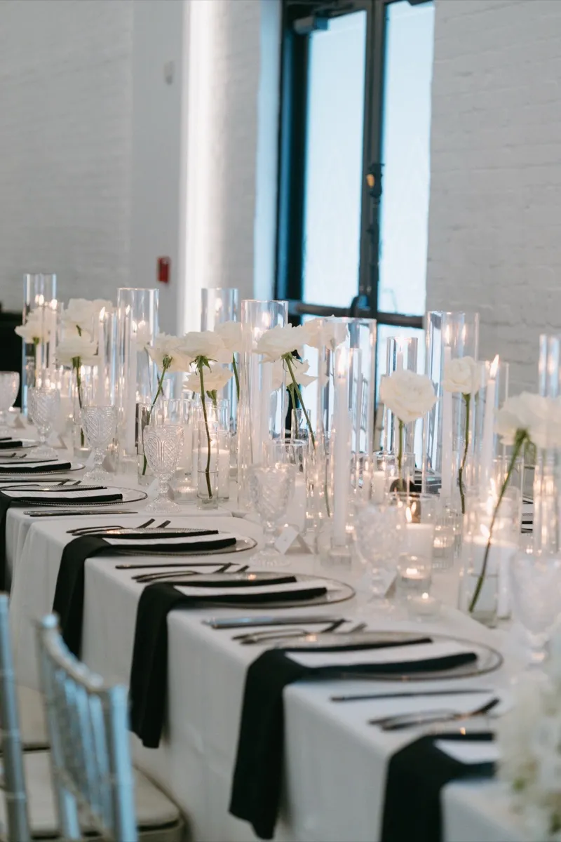 A long reception table set with single white roses in clear bud vases, tall taper candles in glass cylinders, black napkins and cut crystal glassware